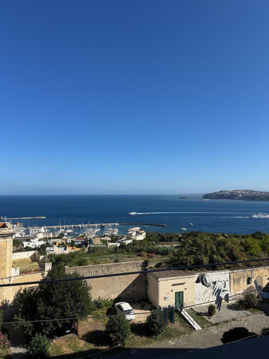 Panoramic view of a Mediterranean coastal town with marina, harbor, and hillside development overlooking clear blue waters.