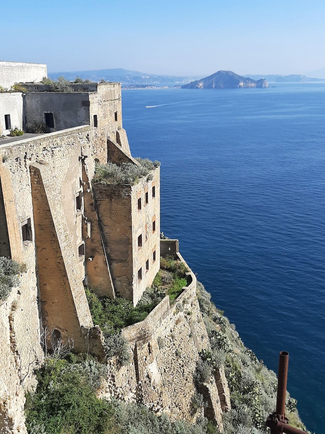 A ruined coastal fortification overlooks the sparkling Mediterranean Sea with distant islands visible on the horizon.