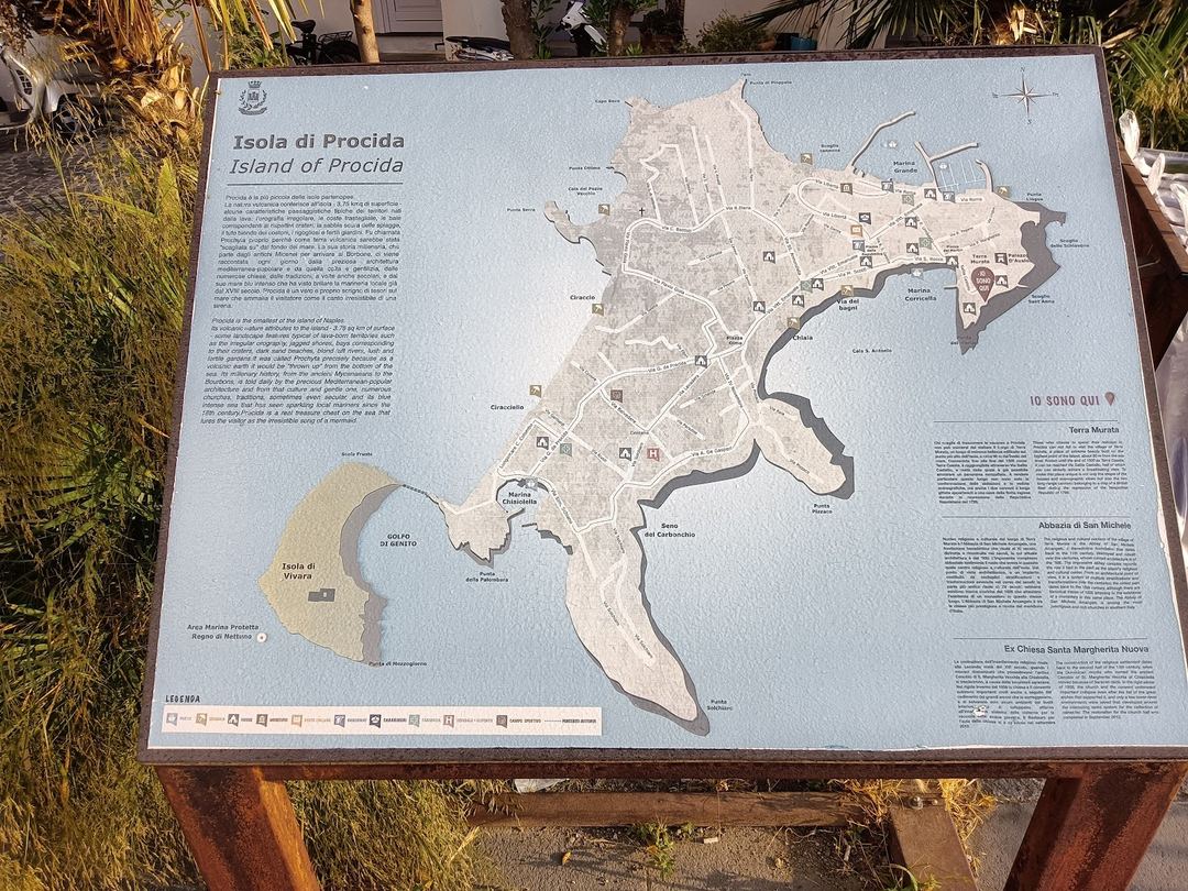 An informational plaque displaying a detailed map of Procida Island with bilingual text in Italian and English, including landmarks and points of interest.
