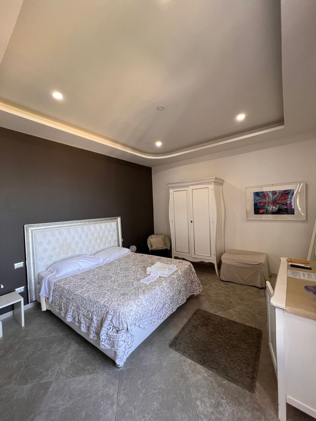 A spacious bedroom with a white upholstered bed, dark accent wall, modern ceiling with ambient lighting, and white wardrobe furniture.