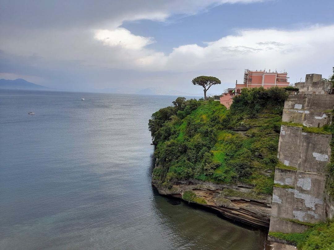 A dramatic coastal clifftop view featuring a distinctive umbrella pine tree and terracotta-roofed building overlooking the Mediterranean Sea with distant mountains on the horizon.