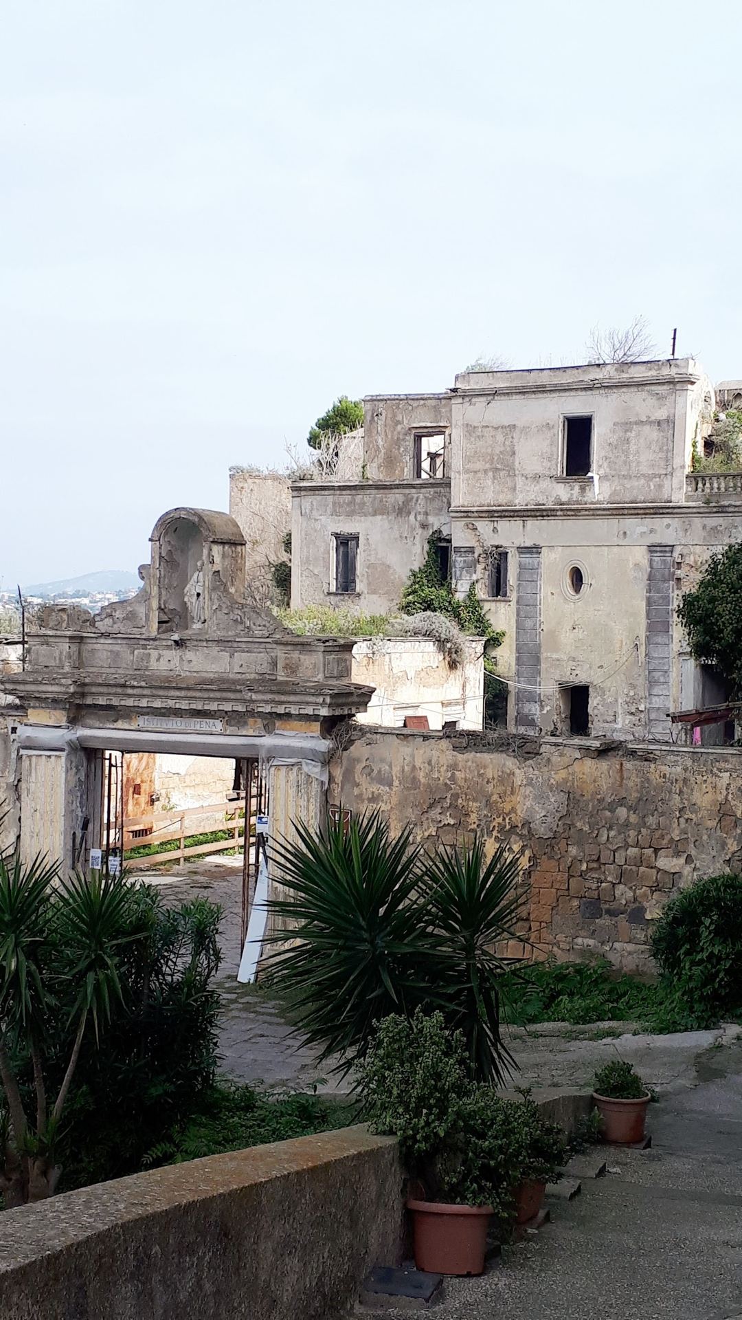 Historic stone villa with Mediterranean architecture featuring weathered stone walls, arched doorway, potted plants, and terraced gardens overlooking the coastal landscape.
